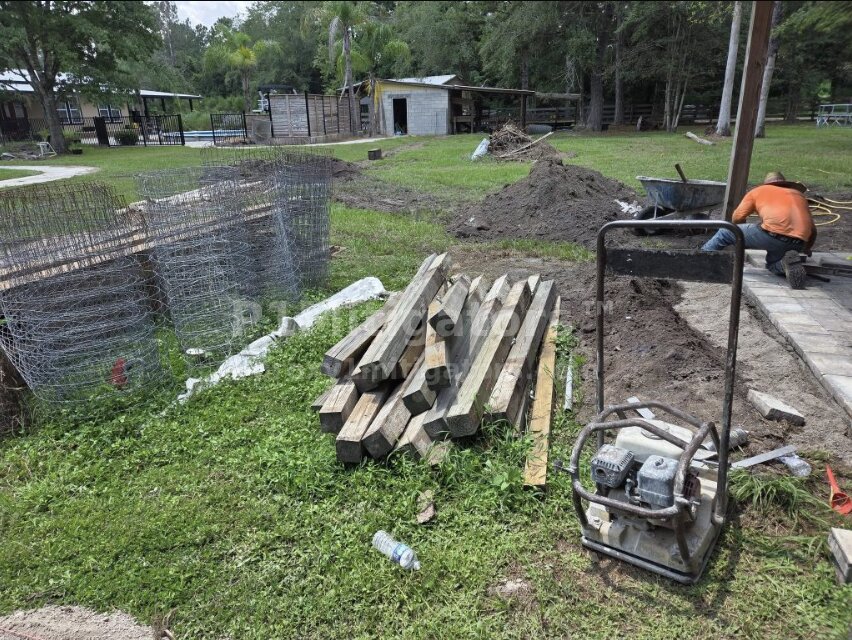 Paver worksite — fence posts, compactor, worker laying interlocking pavers